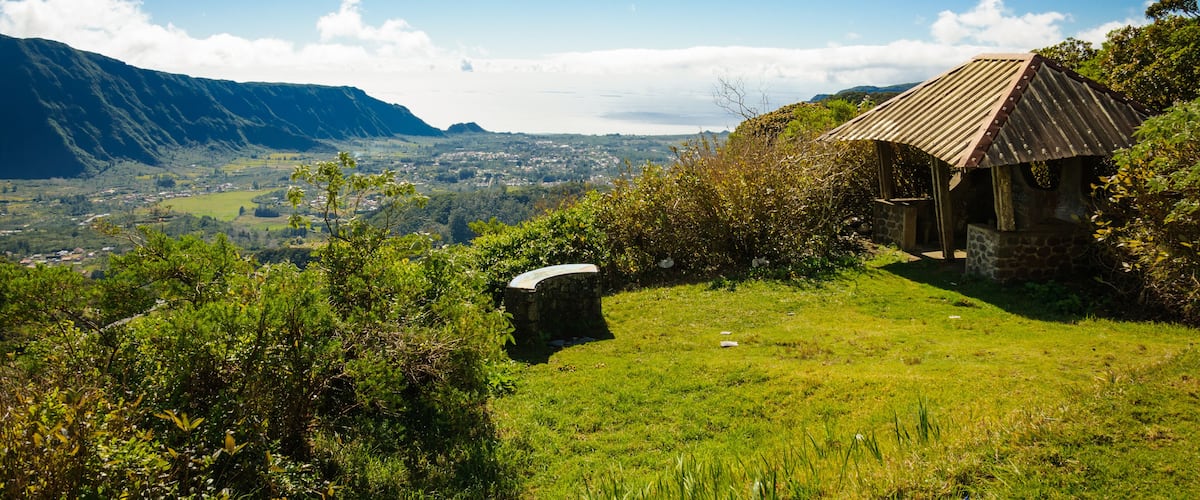 A rural landscape with a stone house, lush greenery, and a distant mountain range under a clear blue sky.