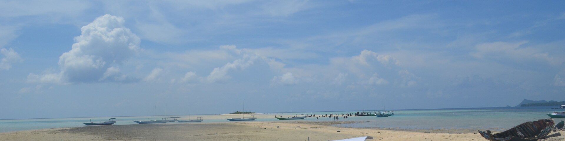 Una playa arenosa con varios barcos, cielo despejado y montañas a lo lejos.