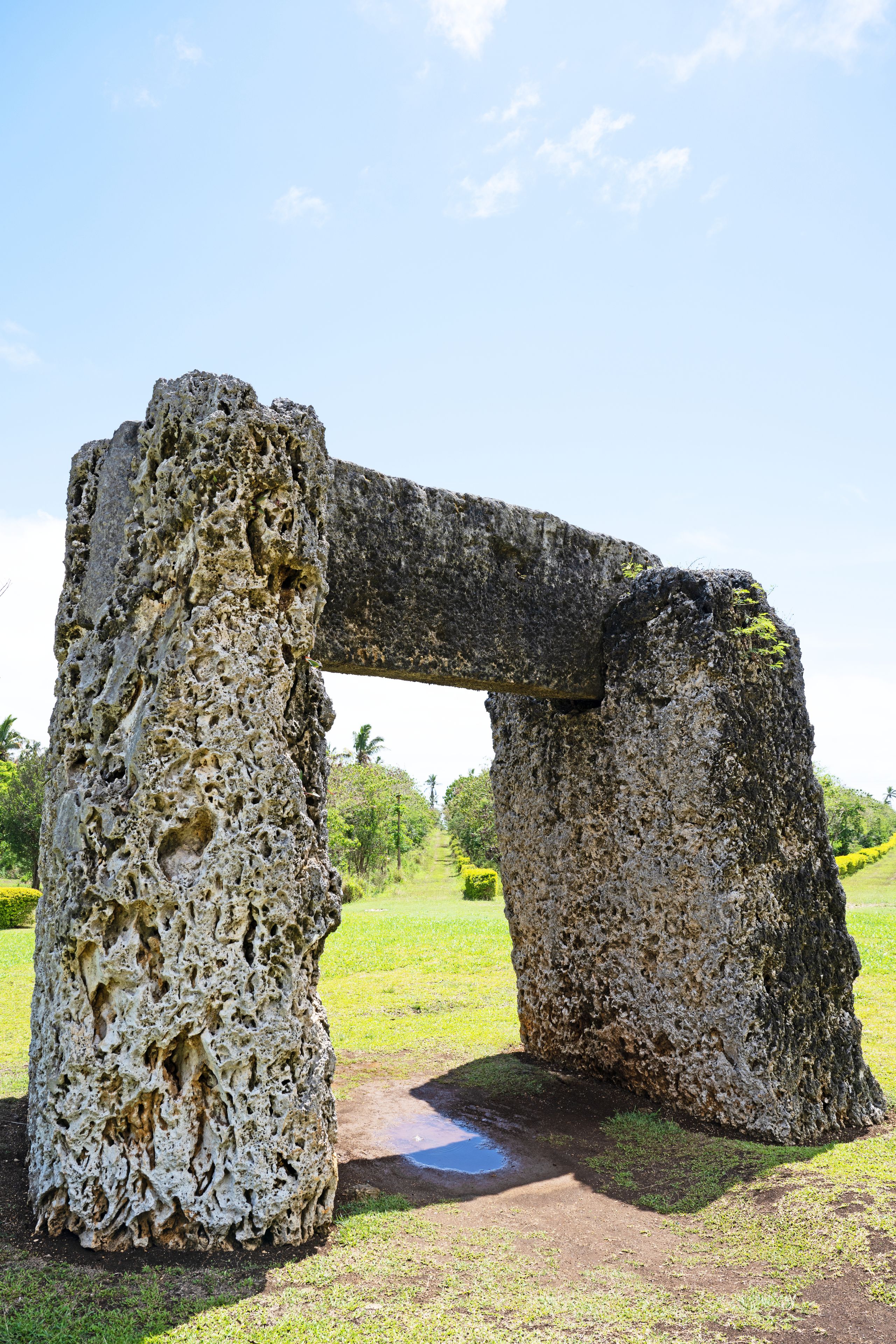 A large, weathered stone archway with intricate carvings, set against a backdrop of a clear sky and a grassy landscape with trees.