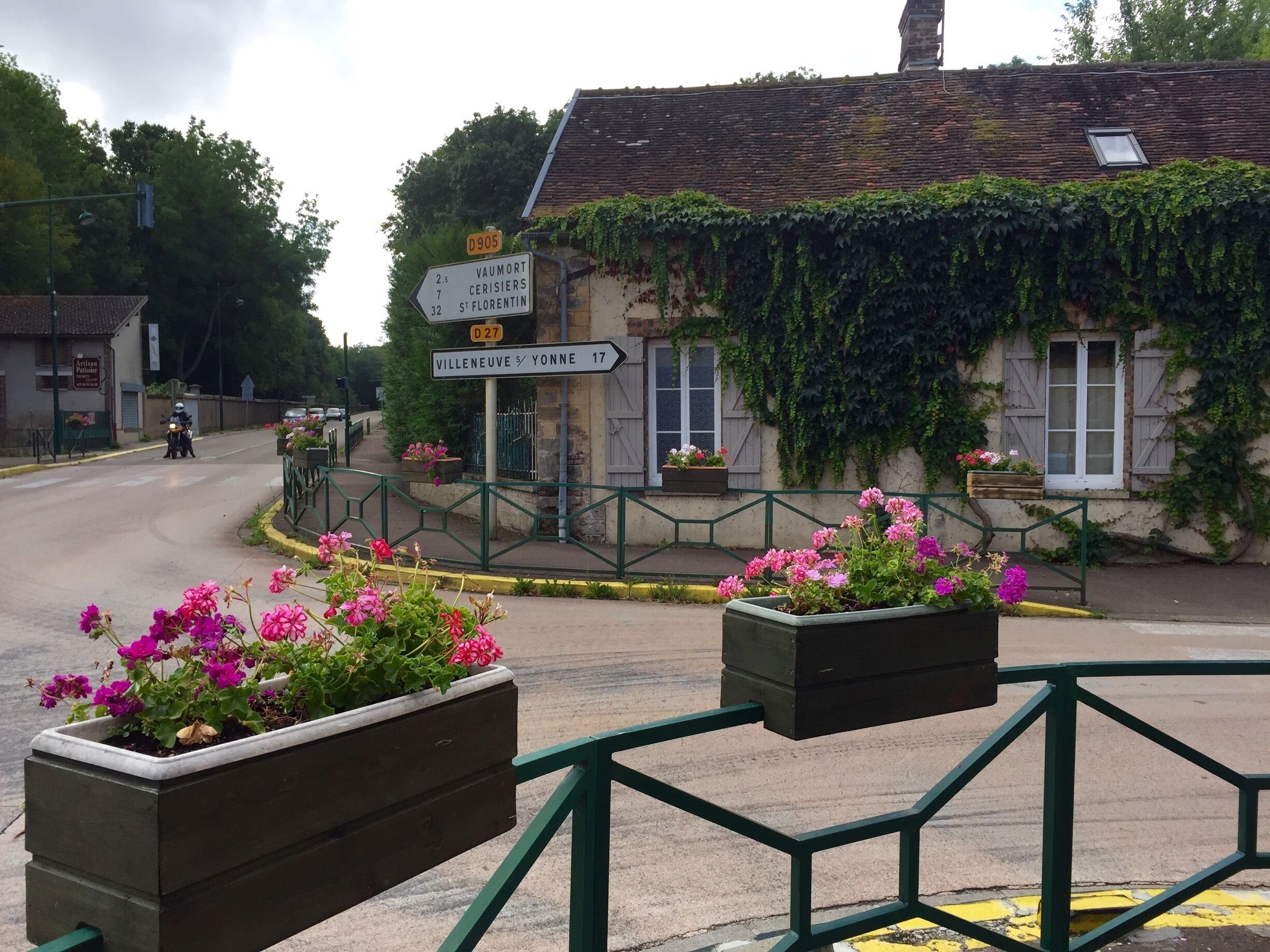 A rural road with directional signs, a building with ivy, and flower planters.