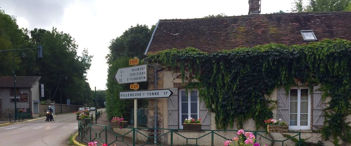 A rural road with directional signs, a building with ivy, and flower planters.