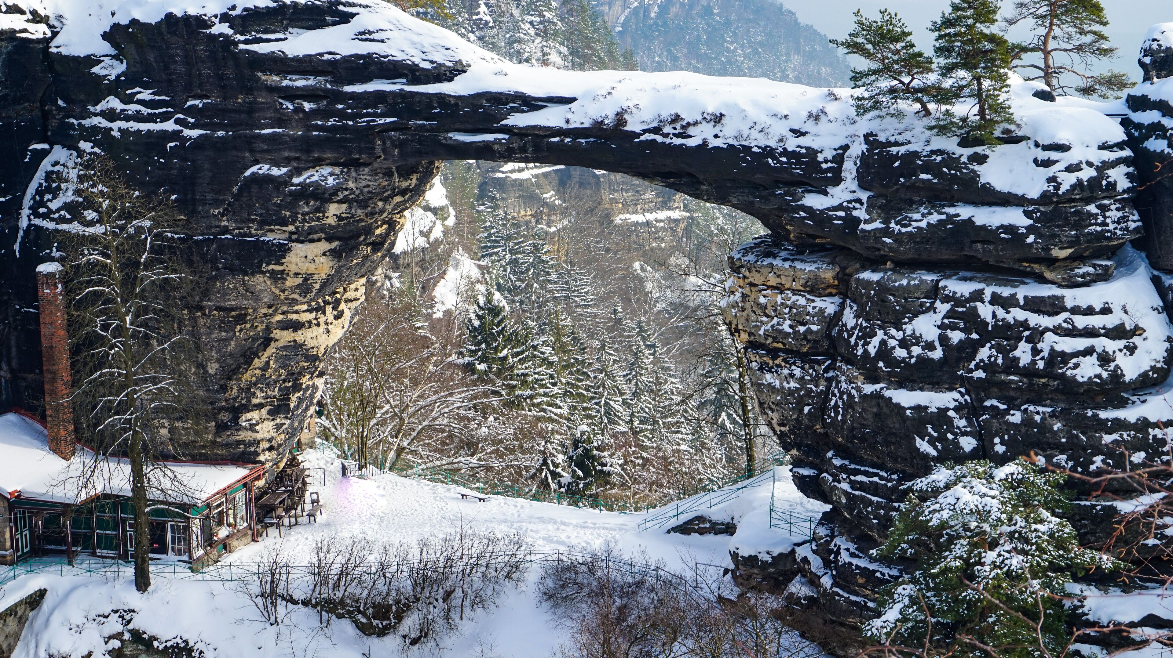 A snow-covered landscape with a large rock arch, trees, and a building with a green roof.