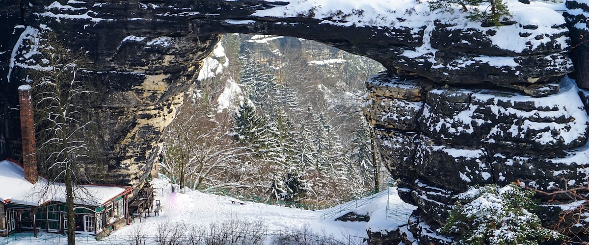 A snow-covered landscape with a large rock arch, trees, and a building with a green roof.