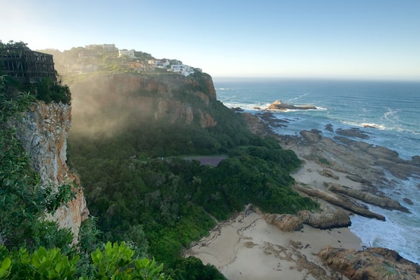 Paisagem costeira com penhasco, praia e edifícios no topo.