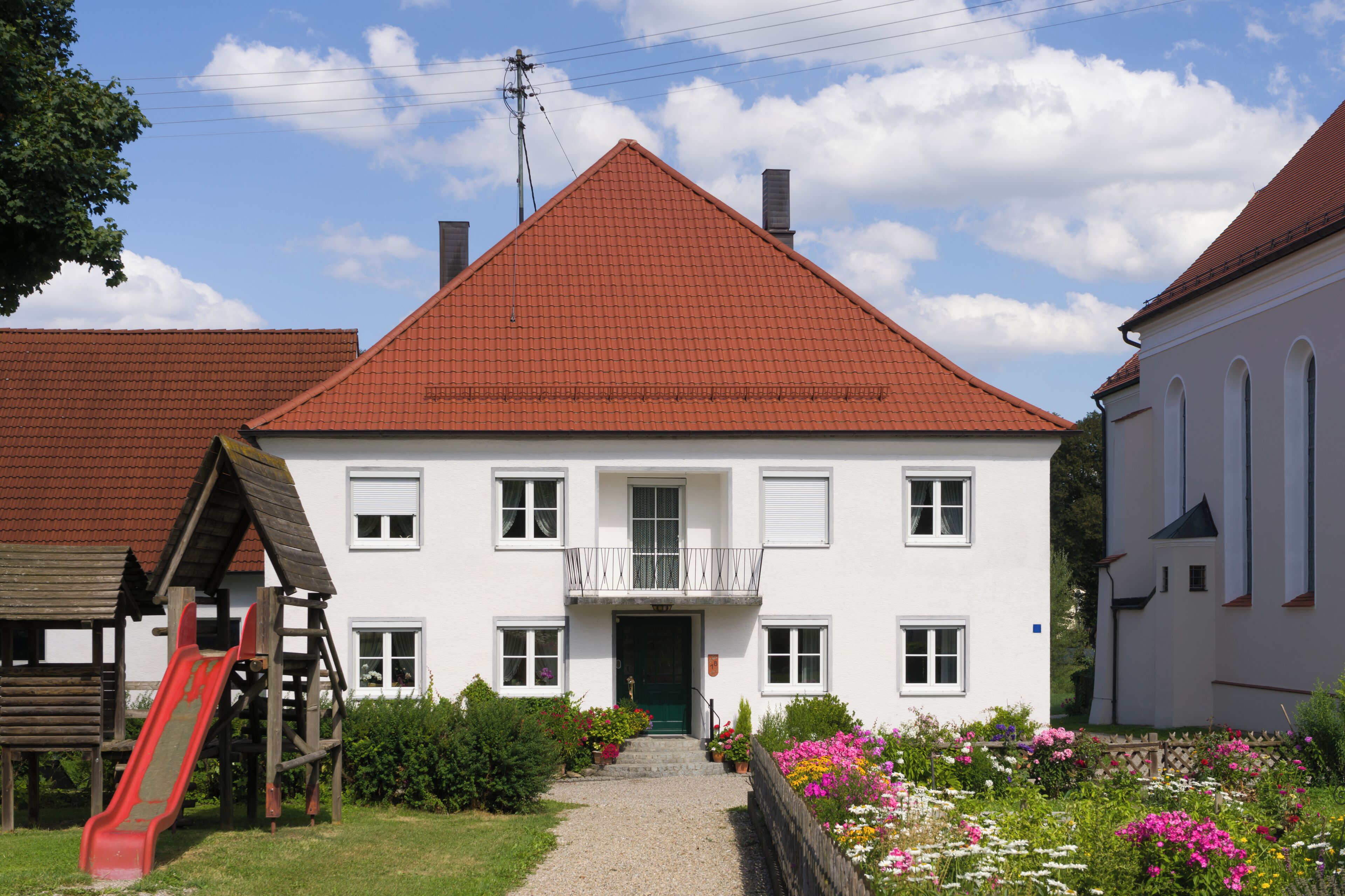 A white building with a red roof, a wooden play structure, and a garden with colorful flowers.