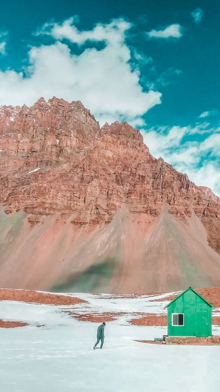 A person walking on snow with a green house and a mountain in the background.