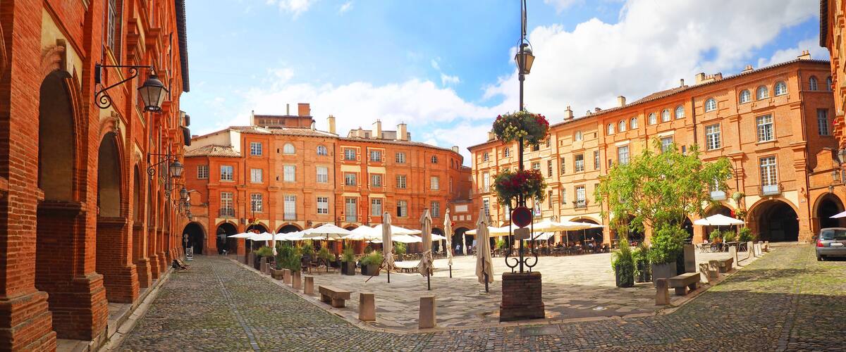 A cobblestone courtyard with red brick buildings, outdoor seating, and hanging flower baskets.