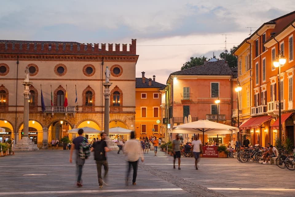 A historic square with a prominent building featuring arched windows and a statue in the center.