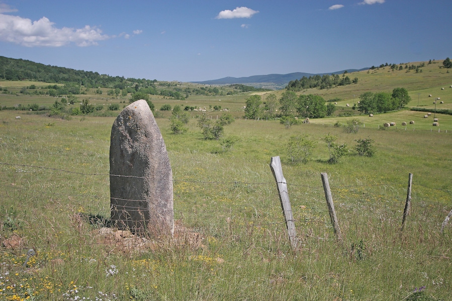 A stone monument in a grassy field with a fence and rolling hills in the background.