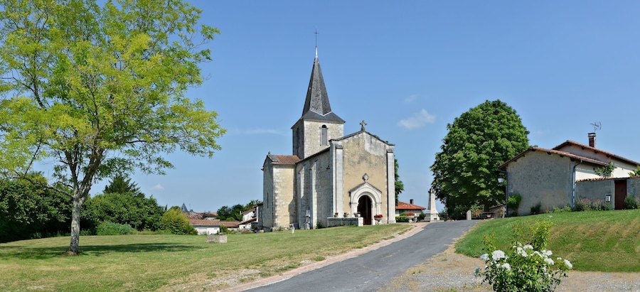 Une église dotée d’un clocher imposant, entourée d’arbres et bordée d’une petite route.