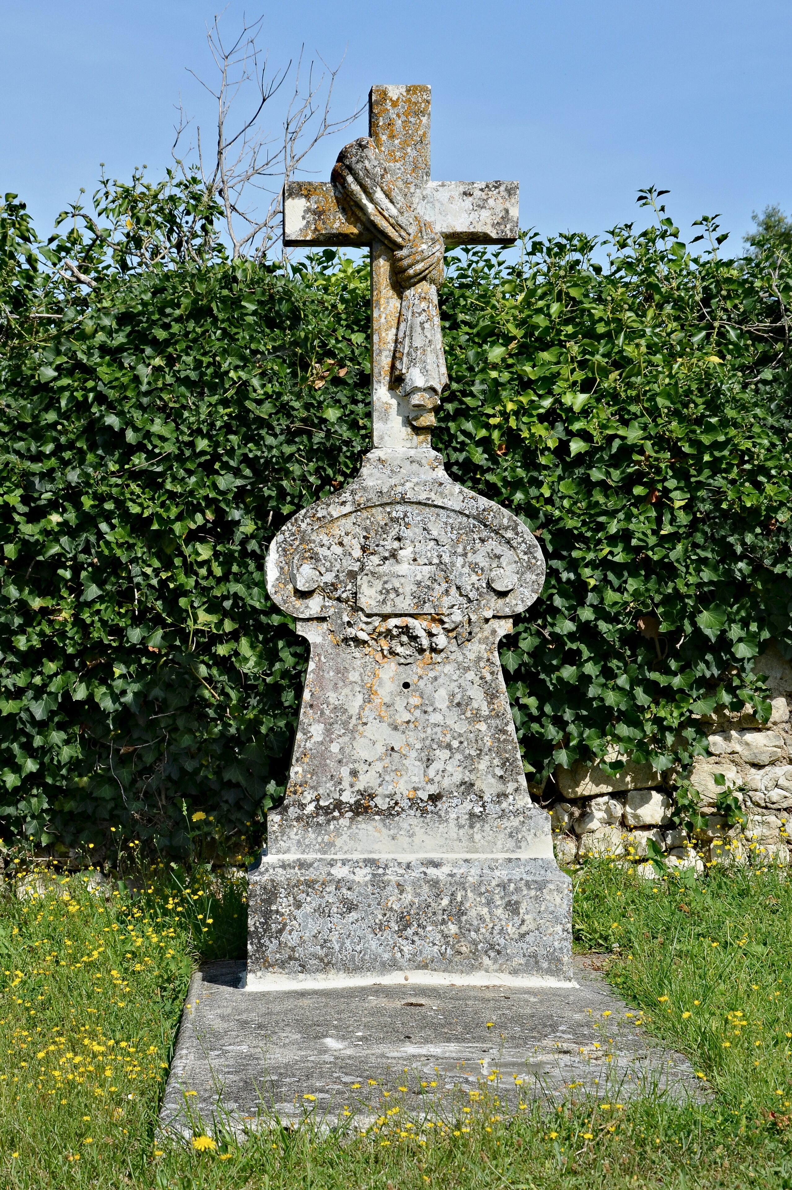 A weathered stone cross with a figure, surrounded by greenery and flowers.