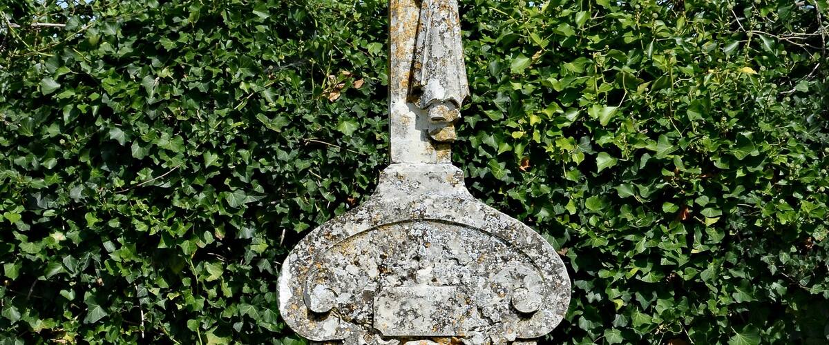 A weathered stone cross with a figure, surrounded by greenery and flowers.