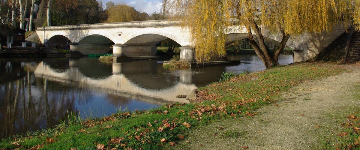 A stone bridge over a calm river, surrounded by trees and a grassy bank.