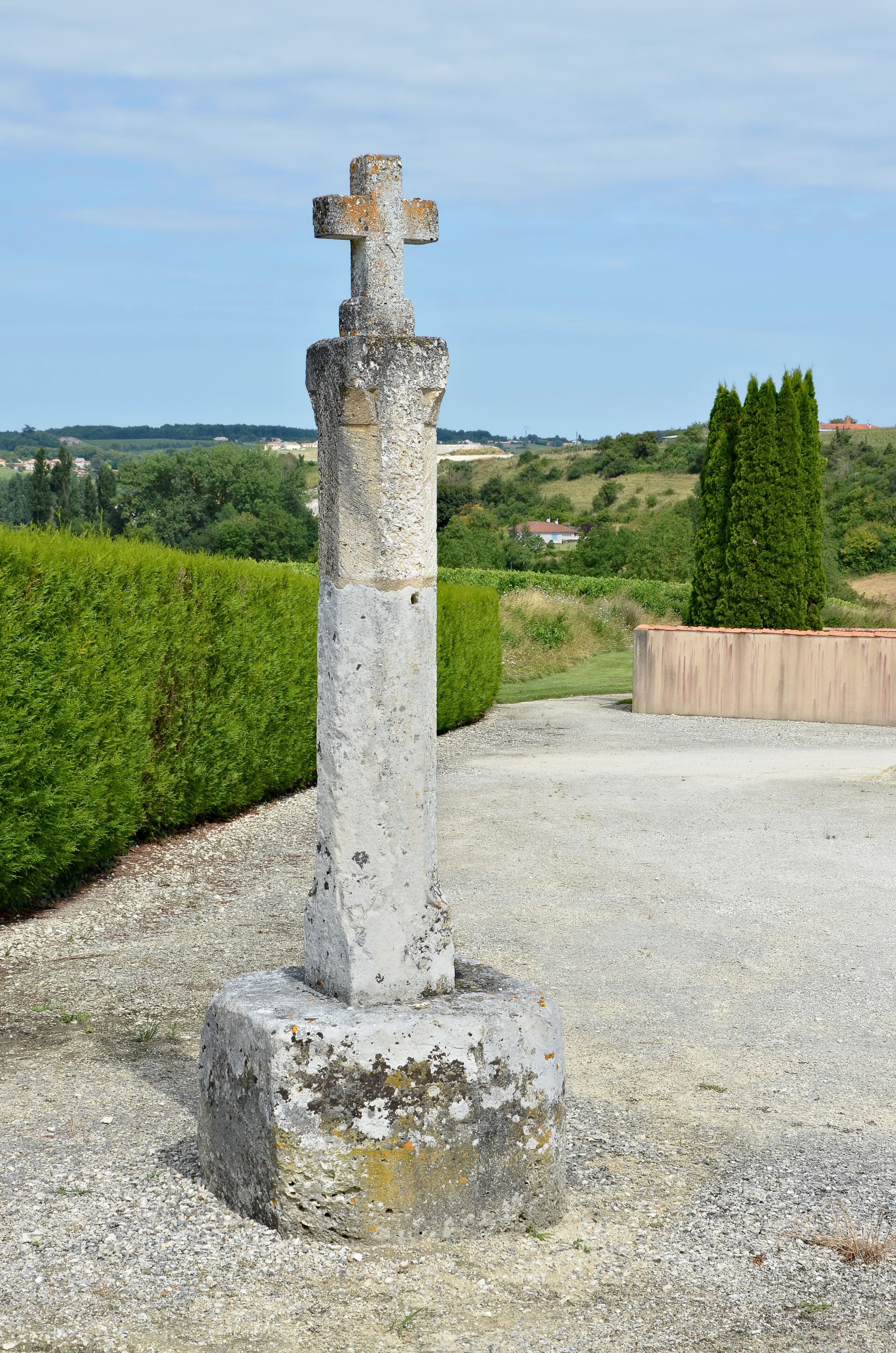 A weathered stone cross in a rural setting with a gravel path, green hedges, and a distant view of houses and trees.