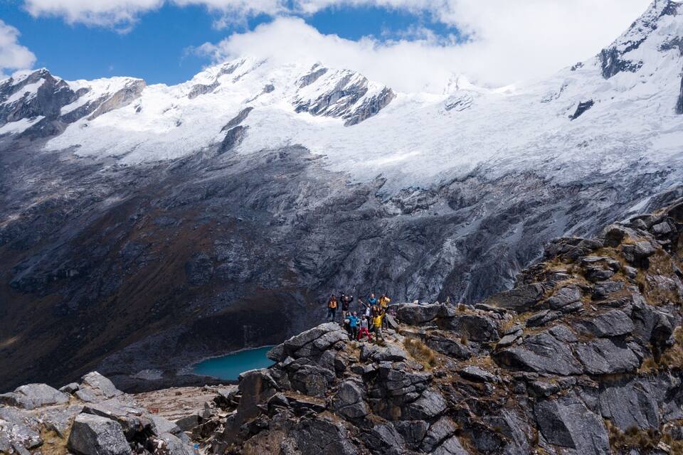A group of hikers on a rocky mountain peak with a turquoise lake below and snow-covered mountains in the background.