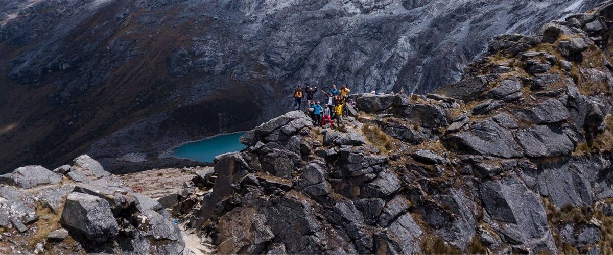 A group of hikers on a rocky mountain peak with a turquoise lake below and snow-covered mountains in the background.