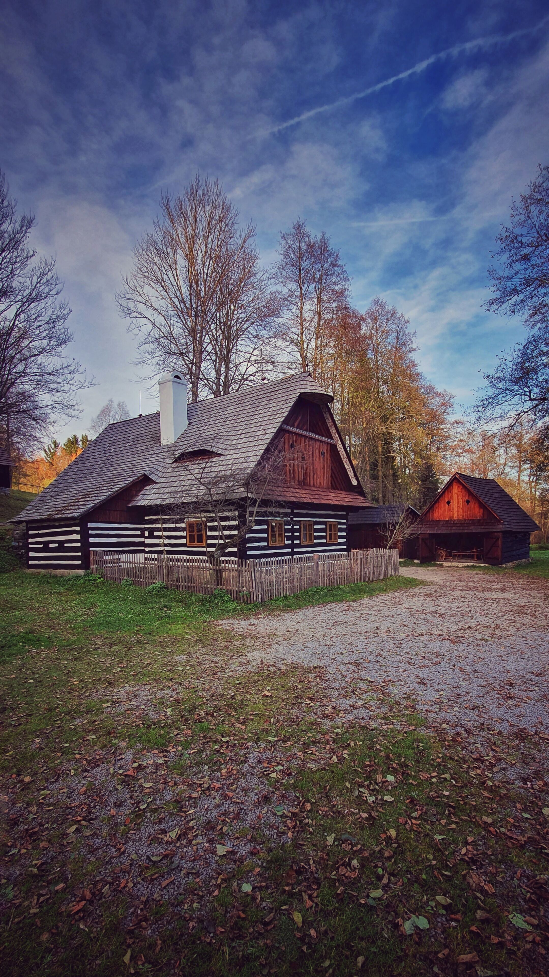 A traditional log cabin with a sloped roof and wooden walls, surrounded by a gravel path and a wooden fence.
