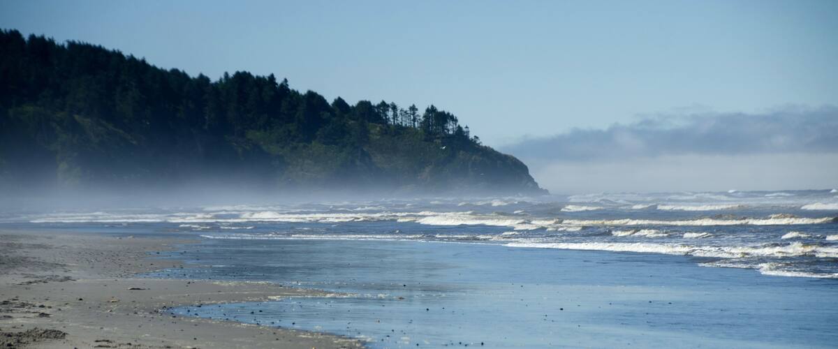 A beach with waves crashing onto the shore, a forested cliff, and a cloudy sky.