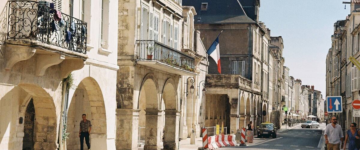 A historic street with stone buildings, a French flag, and a pedestrian crossing.