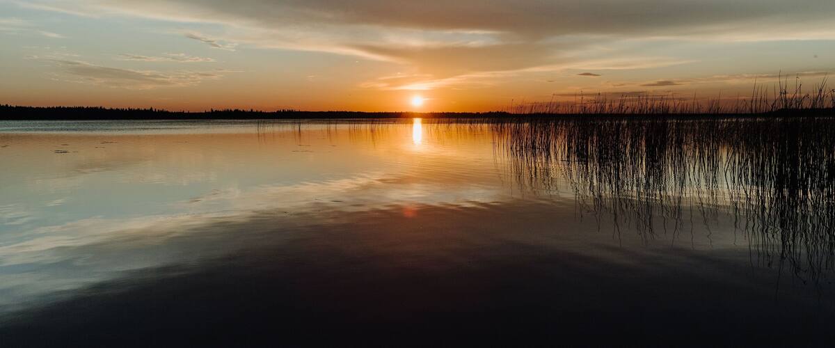 A sunset over a calm lake with reflections of the sky and reeds in the water.
