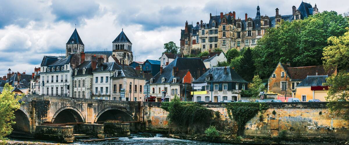 A historic town with a stone bridge over a river, surrounded by colorful buildings and a castle on a hill.