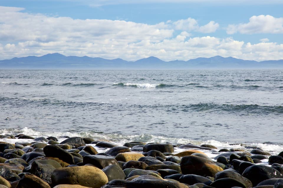 A rocky shoreline with waves crashing onto the rocks, a clear sky, and mountains in the distance.