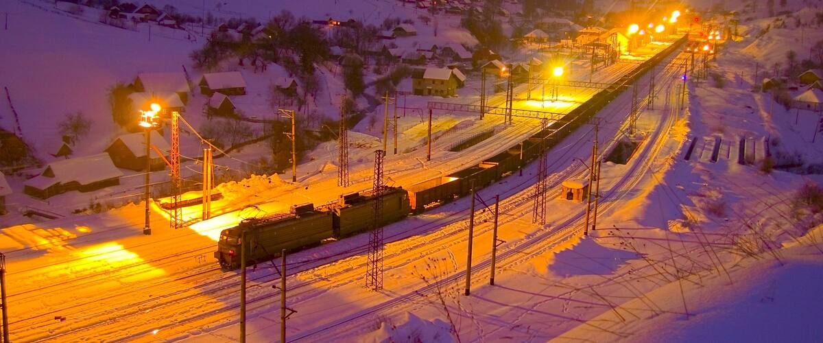A snow-covered railway track with a train and overhead lights in a mountainous area.