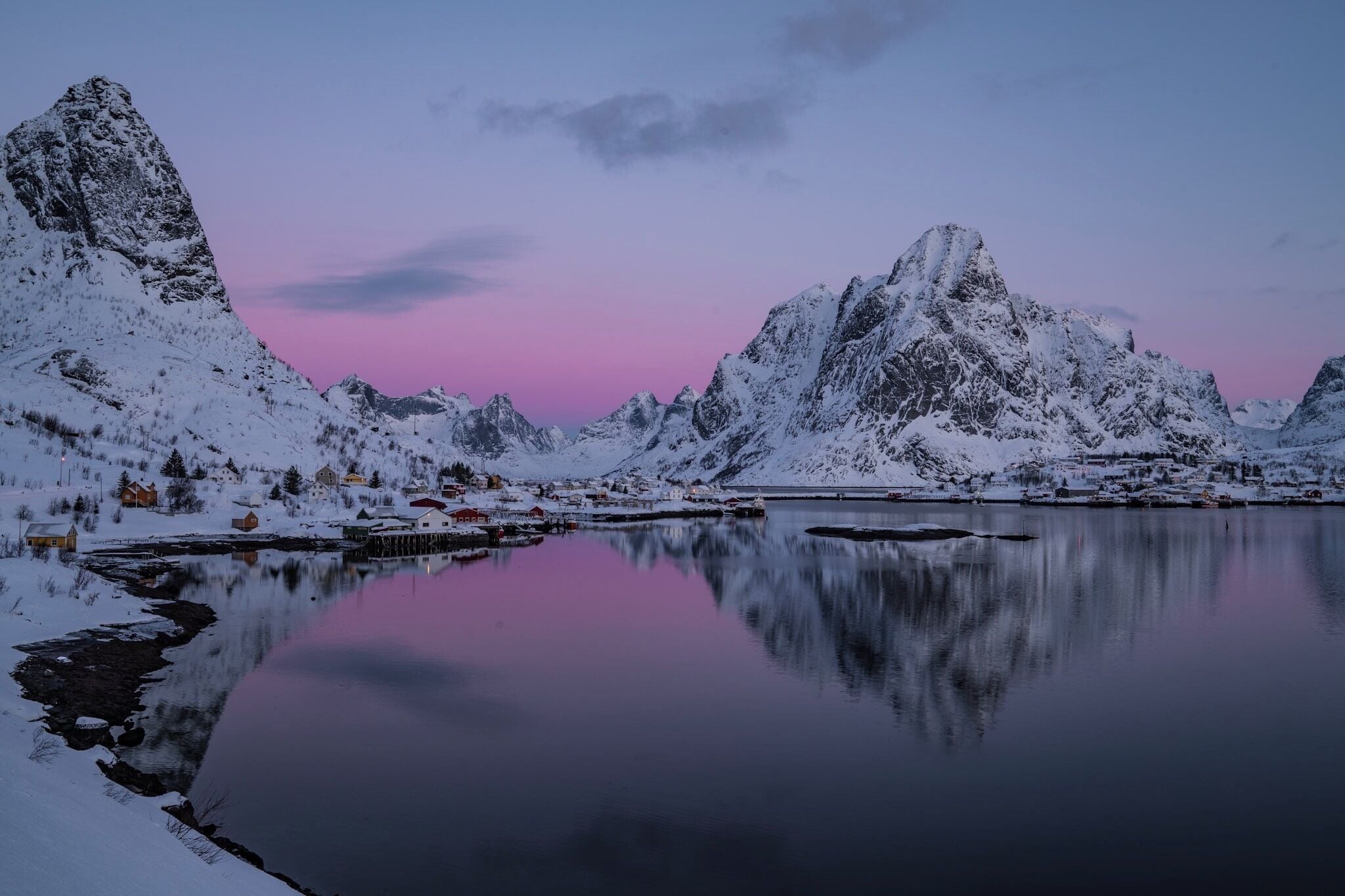 A snow-covered mountain range with a calm lake reflecting the peaks and a pink sky at dusk.