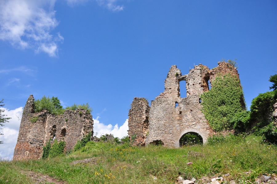 Un château en ruine, fait de pierres, avec des arcades et des tours, entouré de verdure et surplombé d’un ciel bleu clair.