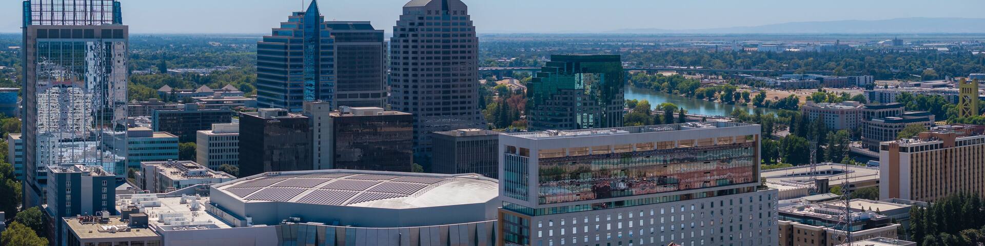 Aerial perspective of Sacramento, California, highlighting the Golden 1 Center, high rise buildings, and the Sacramento River under a clear blue sky.