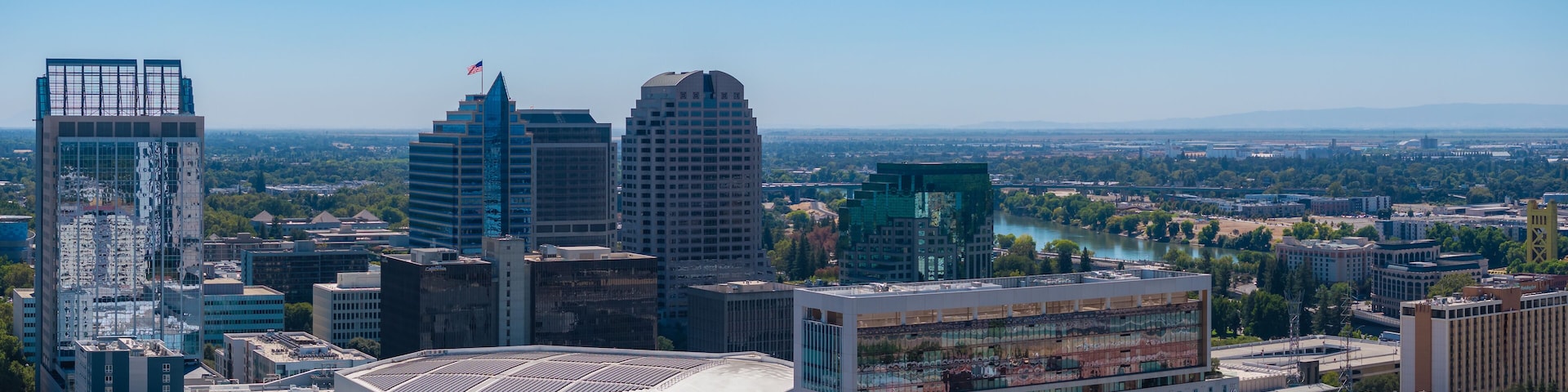 Aerial perspective of Sacramento, California, highlighting the Golden 1 Center, high rise buildings, and the Sacramento River under a clear blue sky.