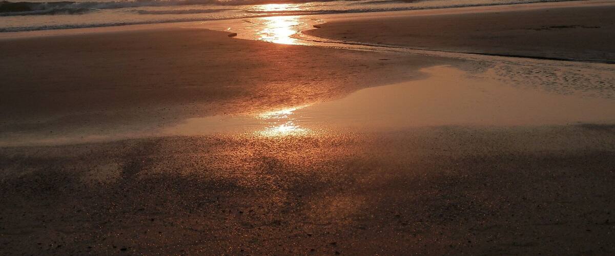 A beach at sunset with the sun reflecting on the wet sand and water.