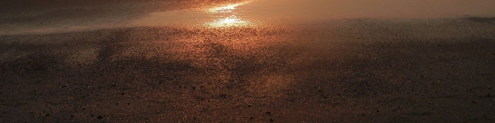 A beach at sunset with the sun reflecting on the wet sand and water.