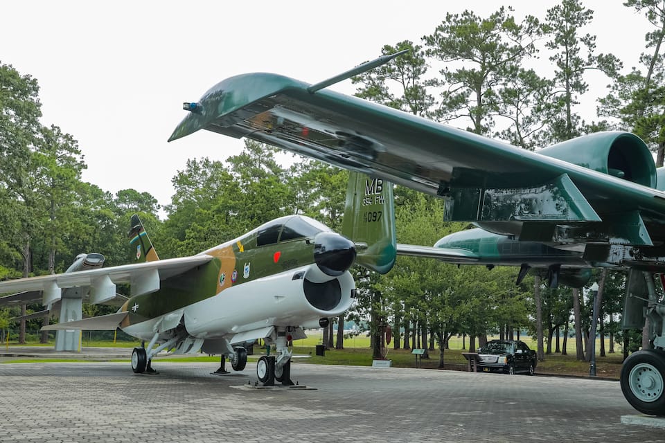 A military aircraft on display, with a green and white camouflage pattern, parked on a paved area surrounded by trees.