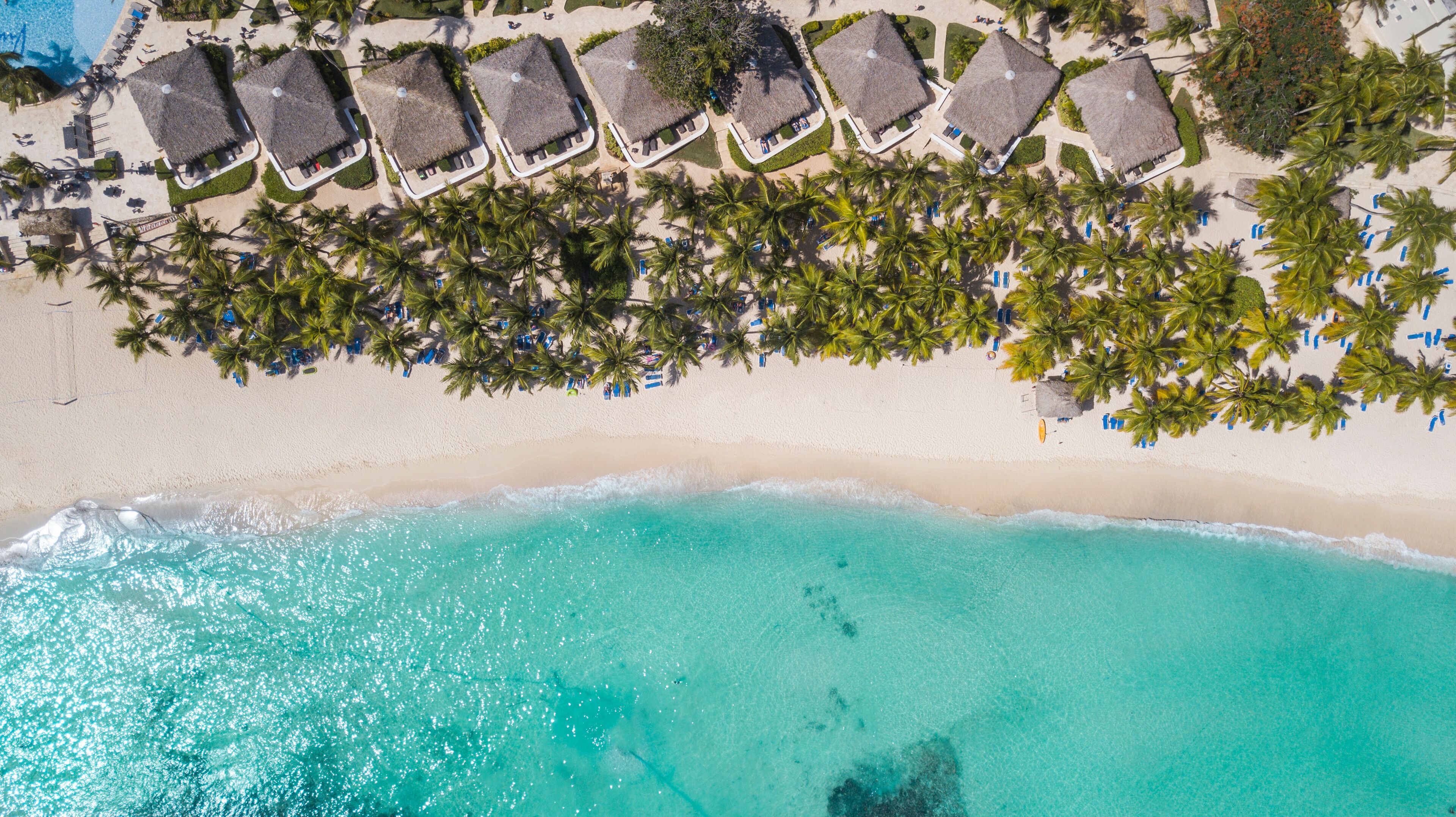 Vista aérea de una playa con aguas turquesas cristalinas, hileras de cabañas de techo de paja y palmeras bordeando la orilla.