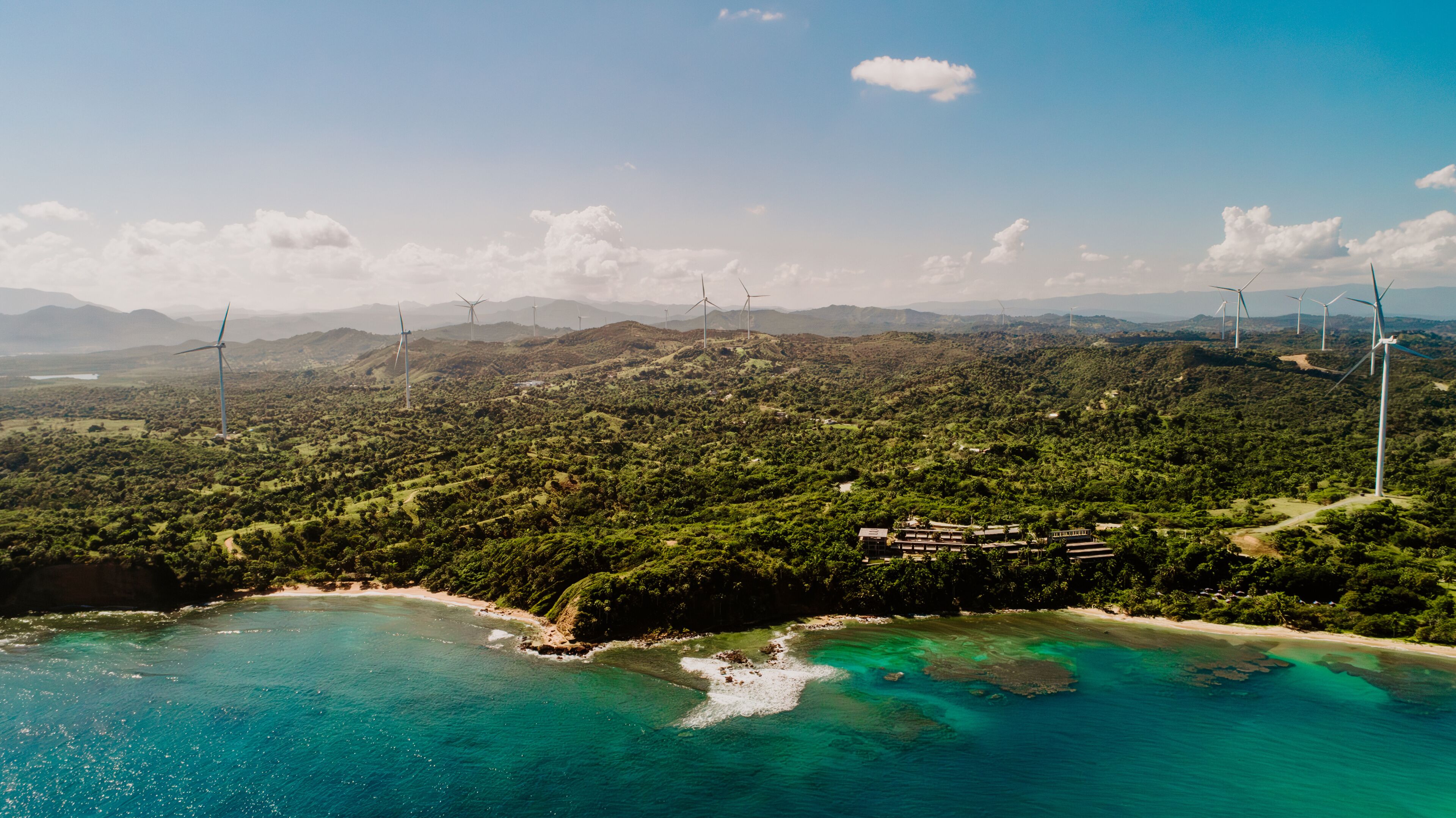 A coastal landscape with wind turbines, a beach, and a mountainous backdrop.