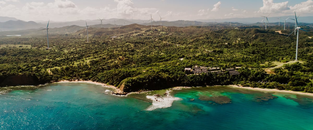A coastal landscape with wind turbines, a beach, and a mountainous backdrop.