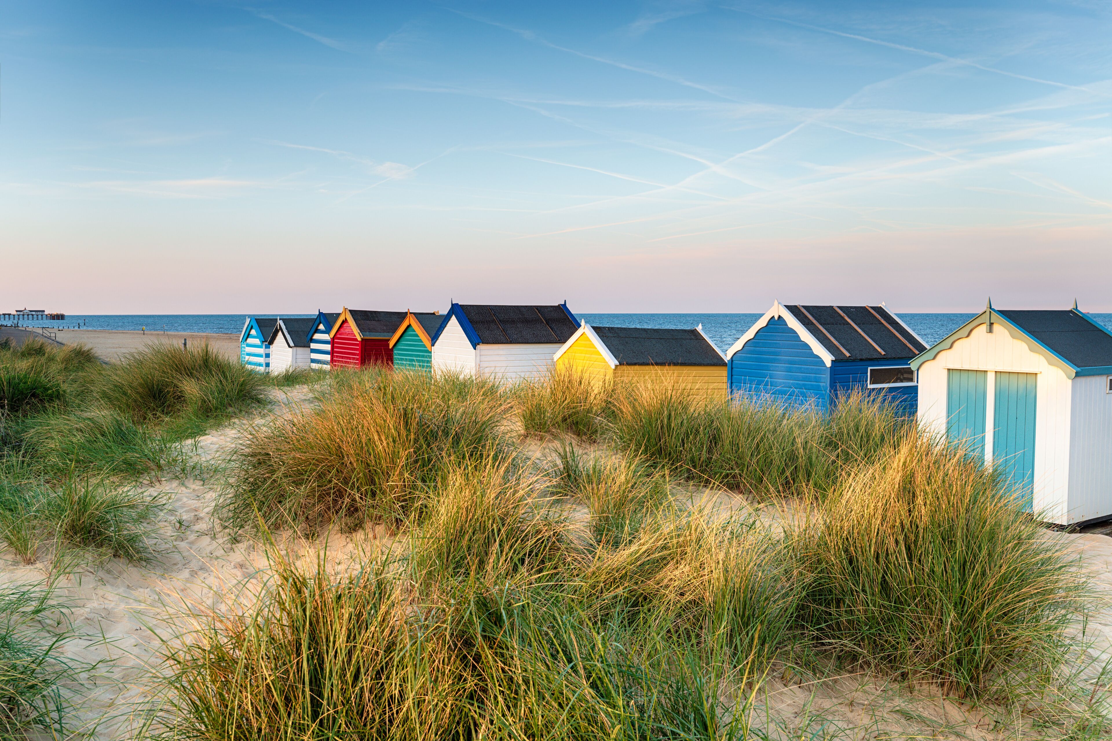 A row of colorful beach huts with a sandy beach and grassy dunes in the foreground.