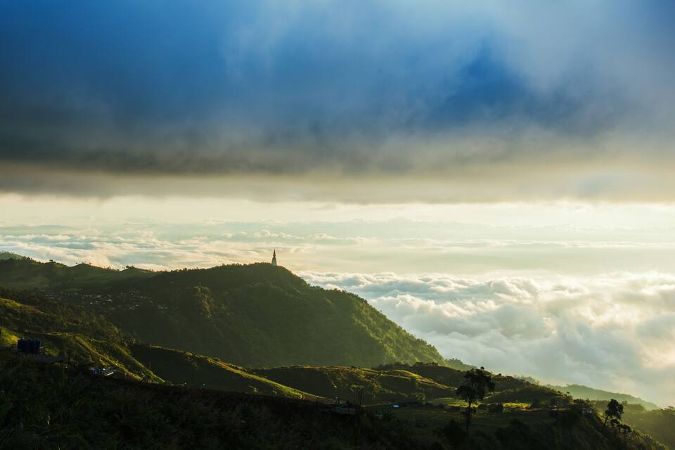 Eine Berglandschaft mit einer Kirche auf einem Hügel, üppigem Grün und einem Wolkenschläng über ihr.