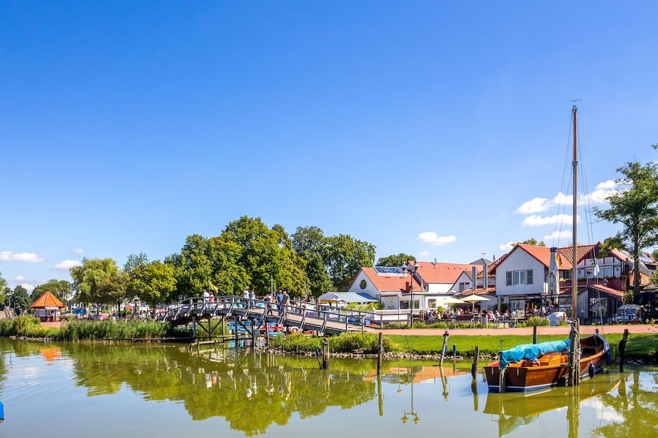 A riverside town with a wooden boat docked near the water, a pedestrian bridge, and buildings with red roofs.