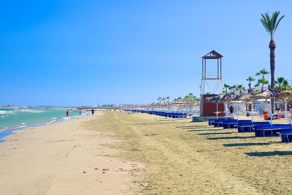 Platja amb cadires blaves per relaxar-se, una torre de socorristes, palmeres i l'oceà.