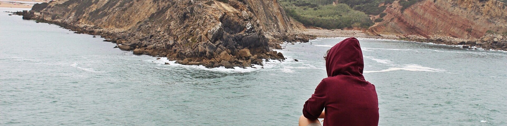 Una persona sentada sobre una roca junto al mar, contemplando un paisaje costero con acantilados y un sendero.