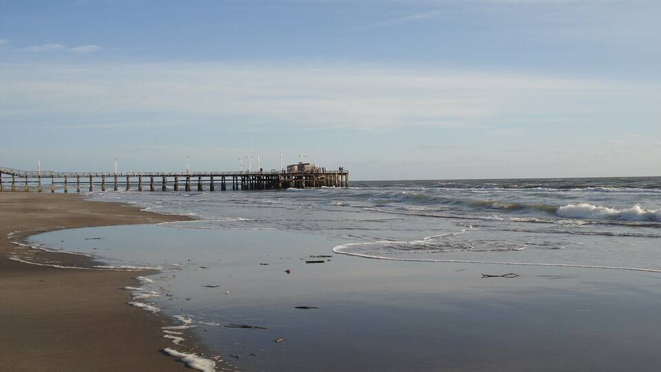 A beach with a pier extending into the ocean, waves crashing onto the shore, and a clear sky.