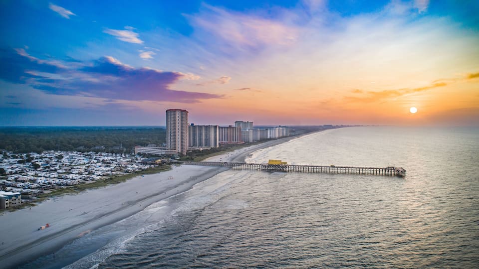 A beach with a pier extending into the ocean, a row of high-rise buildings, and a sunset in the background.