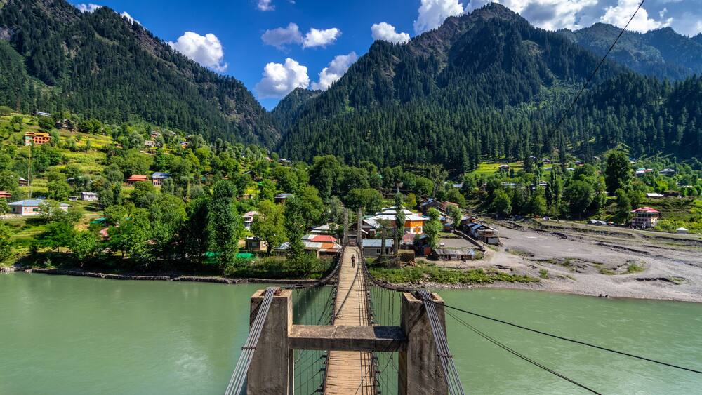View of a suspension bridge leading to a village nestled amidst lush green trees and towering mountains, reflecting in the river, Sharda, Azad Kashmir, Pakistan.