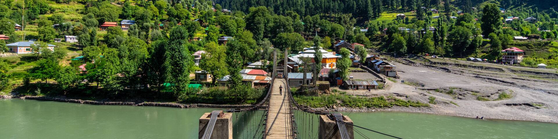 View of a suspension bridge leading to a village nestled amidst lush green trees and towering mountains, reflecting in the river, Sharda, Azad Kashmir, Pakistan.