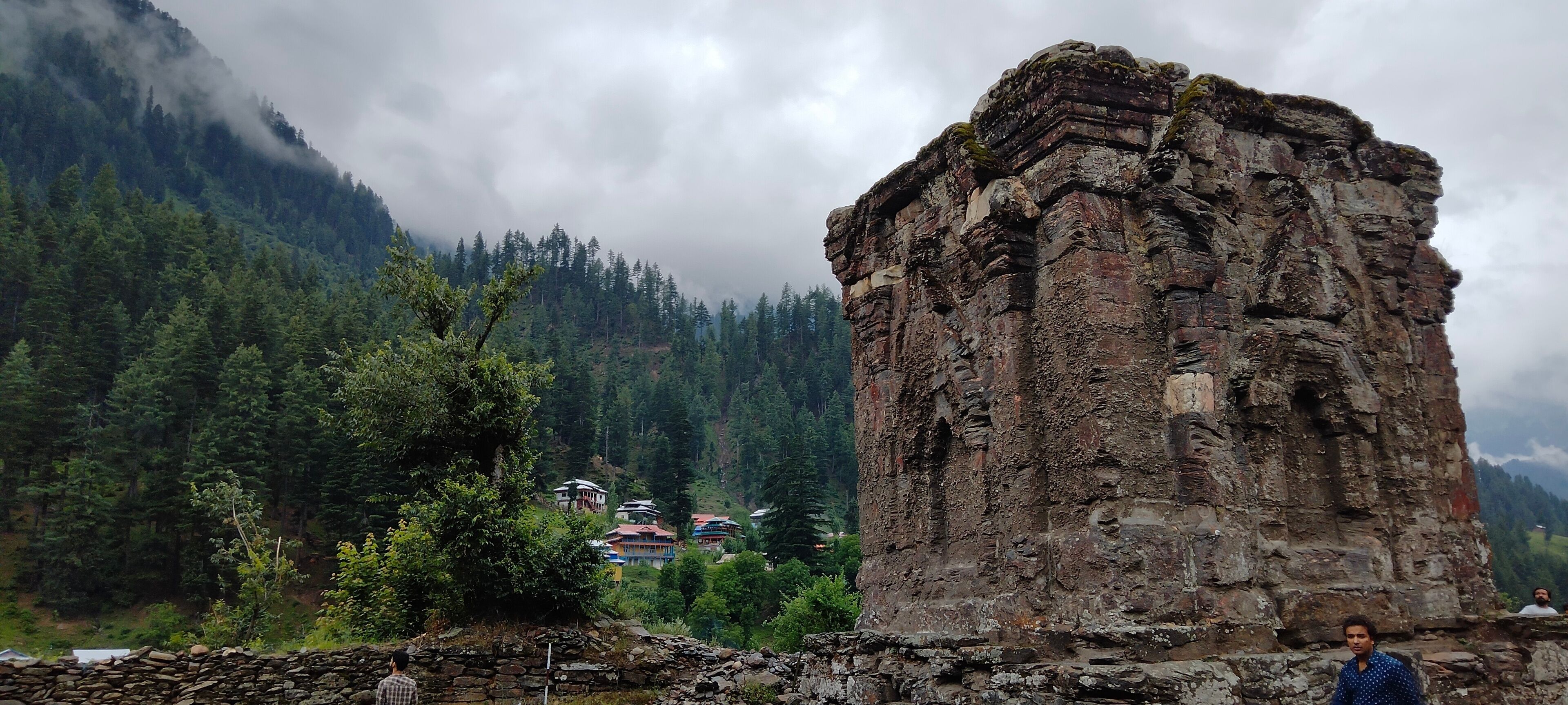 A large stone structure in a mountainous area with dense forests and a few buildings in the background.