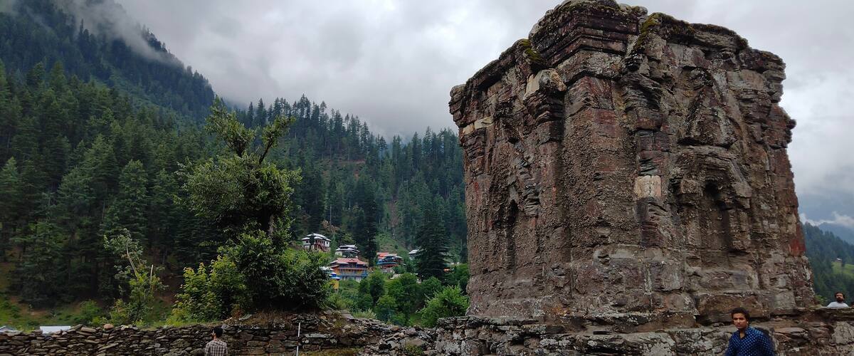 A large stone structure in a mountainous area with dense forests and a few buildings in the background.