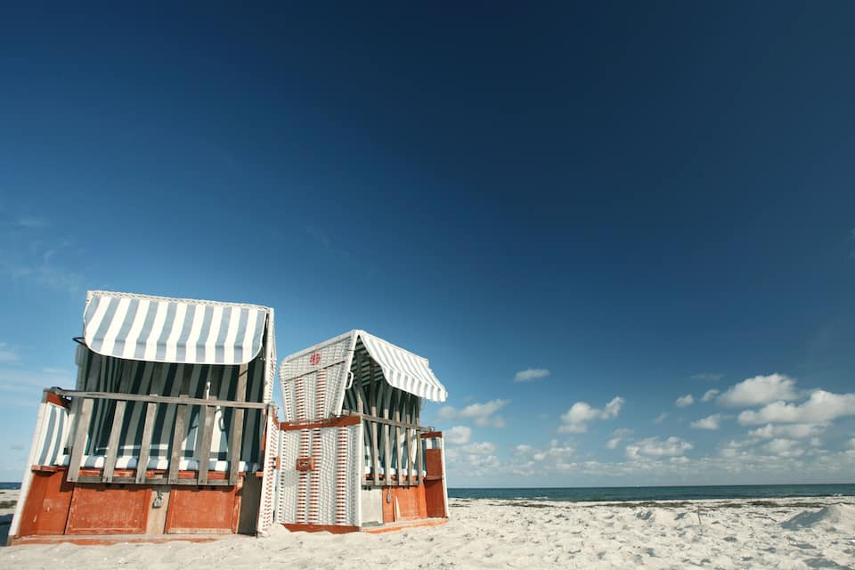 Two beach huts with striped awnings on a sandy beach under a clear blue sky.