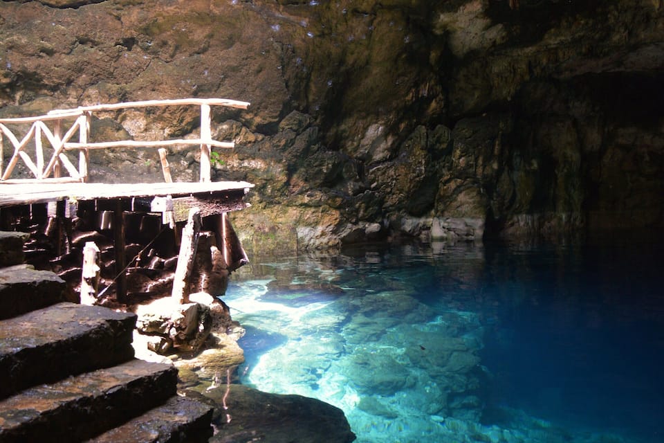 A rocky cave with clear blue water and a wooden staircase leading into it.